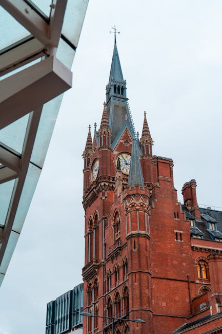 Photograph of a red brick building with a tall, pointed clock tower and ornate architectural details, viewed from beneath a modern building's glass and metal balcony structure. The clock face is visible on the tower, and the sky is overcast. The scene captures a typical cityscape reflecting historic architecture in central London, near St Pancras, with the image framing the tower against the sky. This setting is relevant to house removals and relocation services, illustrating an urban environment where furniture transport and home moving activities might take place, supported by Man with Van St Pancras’s professional moving solutions.