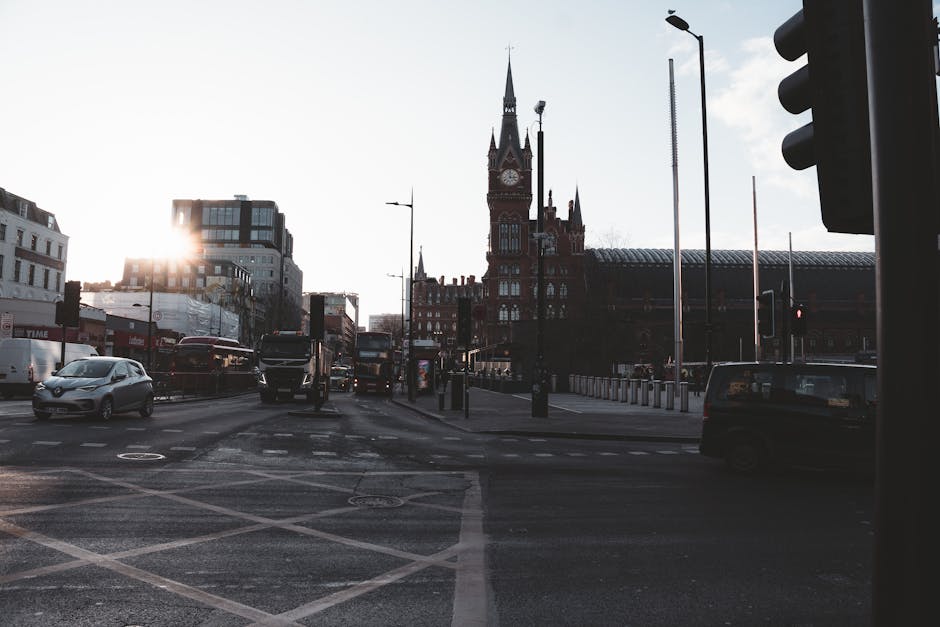 Photograph of a busy urban street scene in St Pancras, London, taken during late afternoon or early evening with the sun setting behind buildings. On the left side, cars and buses are moving through an intersection with clearly marked pedestrian crossings. In the background, there is a historic red-brick Victorian-style station building with a prominent clock tower and spires, partially obscured by the shadow of the surrounding modern structures. To the right, a traffic light pole with multiple signals is visible, along with streetlights and a tall lamppost. The scene depicts an active transportation hub suitable for home relocation or furniture transport logistics, with vehicles potentially being loaded or unloaded in the vicinity. The image overall reflects urban transport activity connected to house and furniture removals, as managed by companies like Man with Van St Pancras.