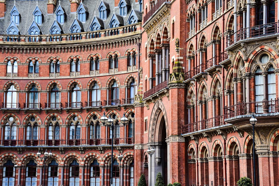 Photograph of a red brick building with a tall, pointed clock tower and ornate architectural details, viewed from beneath a modern building's glass and metal balcony structure. The clock face is visible on the tower, and the sky is overcast. The scene captures a typical cityscape reflecting historic architecture in central London, near St Pancras, with the image framing the tower against the sky. This setting is relevant to house removals and relocation services, illustrating an urban environment where furniture transport and home moving activities might take place, supported by Man with Van St Pancras’s professional moving solutions.