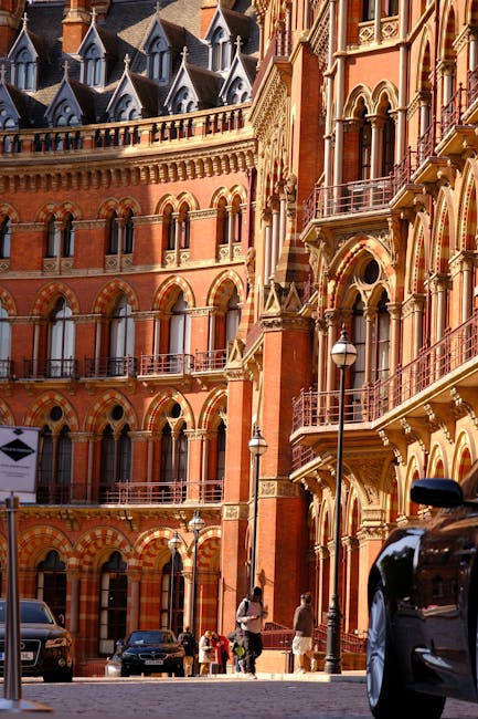 View of a historic, multi-story red brick building with ornate Gothic architectural details, including arched windows and decorative stonework, situated along a paved street in St Pancras. Several pedestrians are walking on the sidewalk, some carrying bags or pushing strollers, while a black van is partially visible in the foreground, possibly involved in a home relocation or furniture transport. Street lamps and parked cars line the street, and the building's balconies and turrets are visible at the upper levels. The scene is lit by natural daylight, highlighting the intricate design and urban environment associated with moving and packing services. Man with Van St Pancras provides relocation expertise, supporting clients with furniture transport, packing, and loading processes within the area.