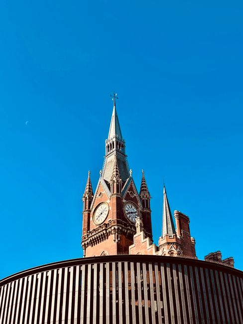 View of a historic, multi-story red brick building with ornate Gothic architectural details, including arched windows and decorative stonework, situated along a paved street in St Pancras. Several pedestrians are walking on the sidewalk, some carrying bags or pushing strollers, while a black van is partially visible in the foreground, possibly involved in a home relocation or furniture transport. Street lamps and parked cars line the street, and the building's balconies and turrets are visible at the upper levels. The scene is lit by natural daylight, highlighting the intricate design and urban environment associated with moving and packing services. Man with Van St Pancras provides relocation expertise, supporting clients with furniture transport, packing, and loading processes within the area.