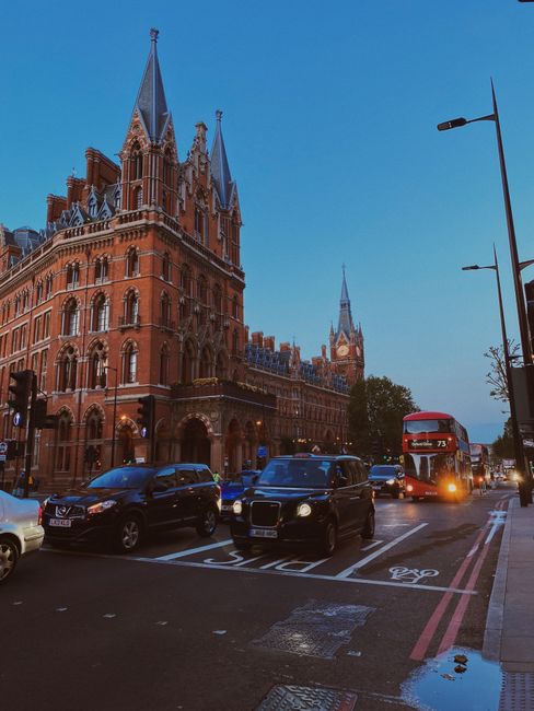 Outside a historic red-brick building with ornate Gothic-style architecture and tall spires, a roadway is busy with traffic including a red double-decker bus, black taxis, and passenger cars. On the pavement, there are parking and bus lane markings, with a street lamp visible on the right side. The scene is illuminated by natural daylight, suggesting a clear sky. This urban environment reflects the area around St Pancras, a prominent landmark in central London. The image captures the typical setting for house removals and moving services, with vehicles and street infrastructure indicating a busy city context where a professional team such as Man with Van St Pancras might operate during a home relocation or furniture transport project.