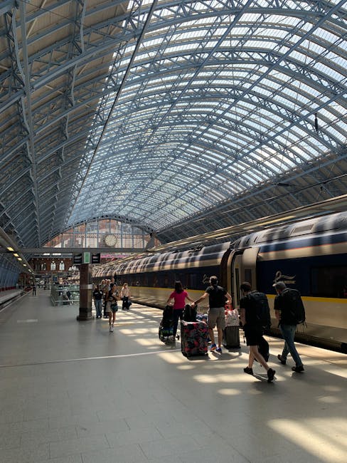 The image depicts the historic St Pancras Railway Station in London, featuring its distinctive red brick facade with ornate architectural details, numerous chimney stacks, and a tall clock tower with a pointed spire. The building’s exterior is illuminated by daylight, with some areas shadowed by cloud cover, showcasing the intricate design elements and large windows typical of Victorian Gothic architecture. In the foreground, a paved public space with a gentle slope and steps is visible, along with a few pedestrians and parked cars near the station entrance. The scene captures the station's prominence as a major transport hub, with its iconic clock tower serving as a focal point, aligning with themes of house removals, home relocation, and furniture transport relevant to [PAGE_TITLE] and the services offered by [COMPANY_NAME].