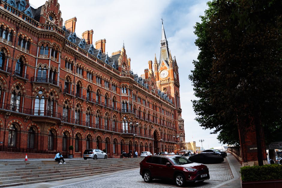 A large, historic red-brick building with ornate architectural details, featuring multiple arched windows with black wrought-iron balconies, situated along a paved street. In front of the building, there are parked cars, including a white van used by Man with Van St Pancras, which is parked near the curb. A person dressed in casual clothing is sitting on the stone steps leading into the building, while others are seen walking nearby. The loading area includes several cardboard boxes wrapped in plastic or brown packing paper, along with wooden furniture pieces and plastic wrap, indicating a home relocation or furniture transport process. Moving blankets, straps, and trolleys are also visible near the van and on the pavement, supporting the loading process. The scene is illuminated by daylight with a partly cloudy sky, and a large tree with green foliage is visible on the right, providing shade over part of the street. This setting reflects typical relocation logistics where packing, loading, and transportation are underway, aligned with house removals services.