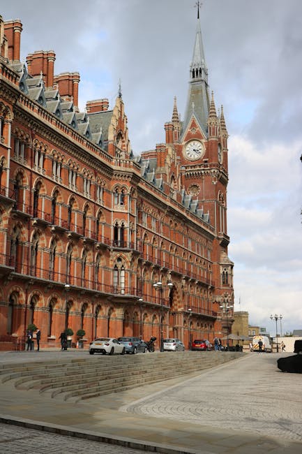 The image depicts a historic red-brick building with ornate architectural details, multiple chimneys, and a prominent clock tower with a spire, situated along a paved promenade. Several cars are parked along the street at the building's base, with some pedestrians walking nearby. The sky above is overcast with grey clouds, providing diffuse natural light over the scene. This setting illustrates a classic urban area suitable for house removals or furniture transport, often involved in residential relocations or packing and moving services. Occasionally, Man with Van St Pancras may use such locations as part of their logistical planning for home relocation projects, ensuring safe and efficient loading and transport of furniture and household items from historic or central locations within London.