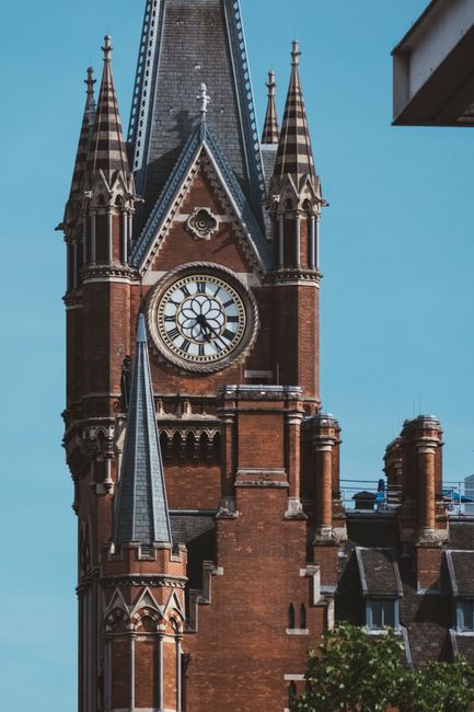 A large, historic red-brick building with ornate architectural details, featuring multiple arched windows with black wrought-iron balconies, situated along a paved street. In front of the building, there are parked cars, including a white van used by Man with Van St Pancras, which is parked near the curb. A person dressed in casual clothing is sitting on the stone steps leading into the building, while others are seen walking nearby. The loading area includes several cardboard boxes wrapped in plastic or brown packing paper, along with wooden furniture pieces and plastic wrap, indicating a home relocation or furniture transport process. Moving blankets, straps, and trolleys are also visible near the van and on the pavement, supporting the loading process. The scene is illuminated by daylight with a partly cloudy sky, and a large tree with green foliage is visible on the right, providing shade over part of the street. This setting reflects typical relocation logistics where packing, loading, and transportation are underway, aligned with house removals services.