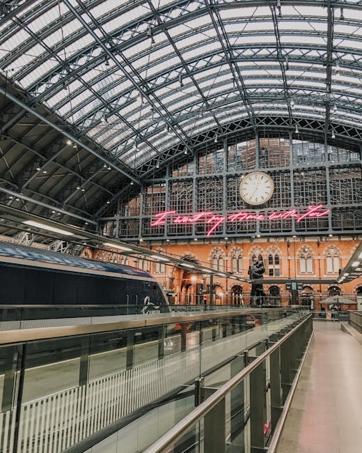 Interior view of St Pancras railway station featuring a high, curved glass and steel roof structure with visible support beams and a large analog clock positioned at the center. Below the clock, there is a bright pink neon sign displaying partially visible text. The station's platform area shows a black train with sleek, modern design parked adjacent to a glass safety barrier with metal handrails. Inside the station, there are numerous cardboard boxes, plastic wrapping, and other packing materials scattered across the platform, indicating ongoing home relocation and furniture transport preparations. A few staff members or movers may be visible loading or organizing the items to facilitate packing and moving services, with a focus on efficient loading process in the context of house removals. The environment is well-lit by natural light filtering through the glass roof, creating a spacious, professional setting suitable for office and household moves, as managed by Man with Van St Pancras.