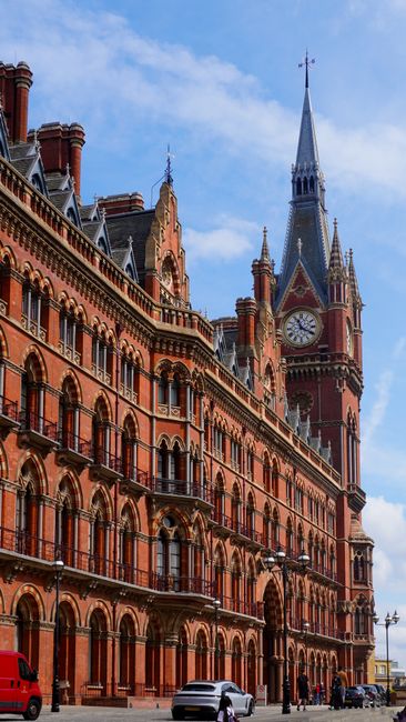 Photograph of a brick building with arched windows and a circular London Underground sign hanging on its exterior wall. Above the building, a historic clock tower with a pointed spire is visible again