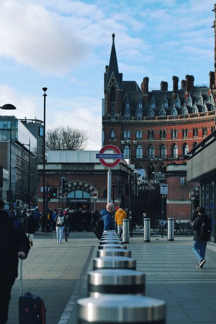 A busy outdoor scene in front of St Pancras train station showing pedestrians walking on the pavement, some carrying luggage or bags. In the foreground, there are metal bicycle racks and a row of boll
