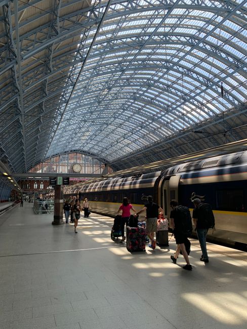 A busy outdoor scene in front of St Pancras train station showing pedestrians walking on the pavement, some carrying luggage or bags. In the foreground, there are metal bicycle racks and a row of boll