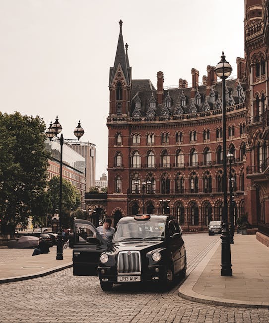 A black London taxi with the license plate LR13 AUP is parked on a cobbled street outside a historic red-brick building in central London, near St Pancras. The taxi's front door is open, and a person in a white shirt is seen unloading or preparing to load items, which could include moving boxes or furniture for a house removal. The scene is illuminated by traditional black lamp posts lining the street, and there are other vehicles visible in the background. Trees are along the sidewalk, and modern office buildings are visible further down the street. This setting illustrates the logistical process of furniture transport and home relocation in an urban environment, with [COMPANY_NAME], Man with Van St Pancras, providing professional removals and packing services nearby for local house moves and student move-outs, as referenced by the page title.