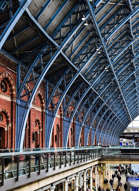 Interior view of St Pancras railway station showcasing its ornate iron and glass vaulted ceiling supported by blue-painted steel arches. The station’s brickwork with arched windows is visible on the upper level, and the lower level features a busy walkway with numerous people walking and standing. Inside the station, a row of luggage trolleys and some scattered cardboard boxes are positioned near the glass railing along the upper platform, indicating ongoing home relocation or furniture transport preparations. The lighting highlights the architectural details of the historic station, with natural light streaming through the large windows. This scene captures the environment where house removals and moving services may encompass loading and unloading activities within a busy transport hub, with Man with Van St Pancras providing professional relocation support in such bustling environments.