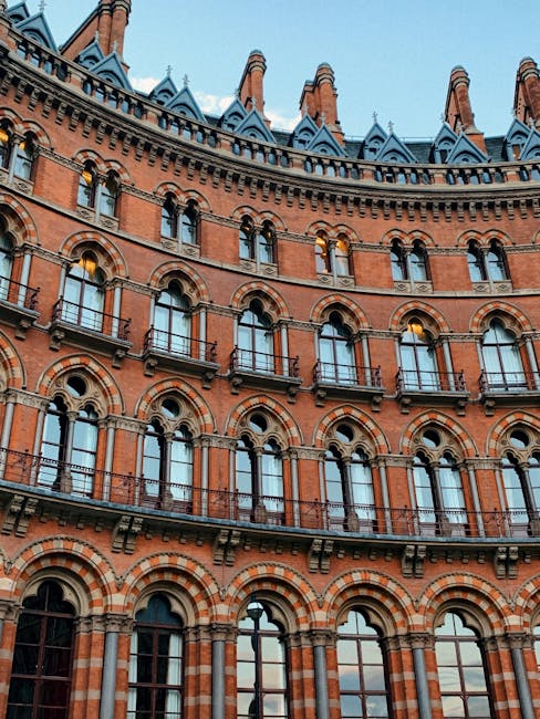 Photograph of a historic red-brick building with ornate architectural details, including arched windows and decorative brickwork, seen from a low angle against a clear sky. The building features multiple stories with black metal balconies and intricate cornices. This backdrop highlights the challenging nature of navigating narrow Georgian stairs during a home relocation. In a separate scene, inside a property, there are cardboard boxes, wrapped furniture with blankets and plastic wrap, and a wooden chair visible near a doorway. A professional moving service, such as Man with Van St Pancras, might be involved in packing and moving fragile items, with equipment like trolleys, straps, and a moving van partially visible outside the building, ready for the furniture transport process. The lighting is natural, emphasizing the historic character of the exterior and the details necessary for efficient house removals in tight staircases.