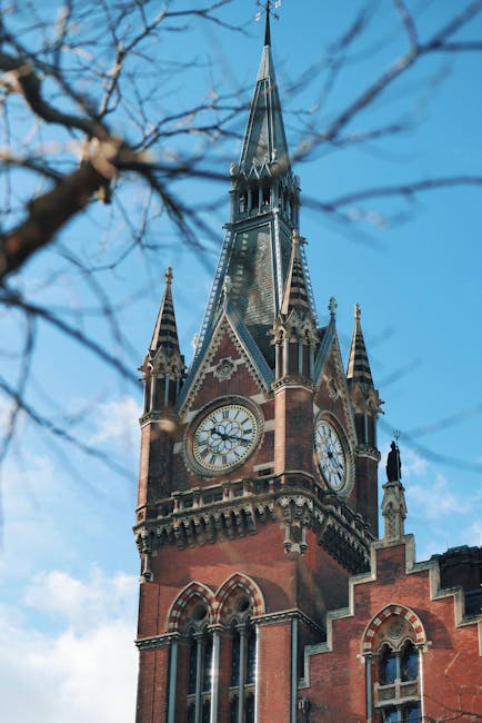 A London Underground sign with a blue and white roundel, mounted on a metal pole in front of a historic red-brick building featuring Gothic architectural elements and a clock tower with pointed spires