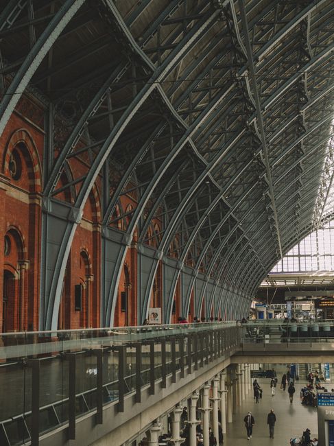 Interior view of a large train station with a high, curved glass and steel roof structure supported by dark metal arches. Red brick walls with arched windows line the upper part of the station, while the lower level features a spacious, open area with a few passengers walking and waiting near ticket booths and information signs. The station's modern glass facade allows natural light to illuminate the space, highlighting the combination of historical brickwork and contemporary architectural elements. The scene depicts a busy transportation hub, suitable for home relocation or furniture transport services like those offered by Man with Van St Pancras, with a focus on the station’s structural design and passenger activity.